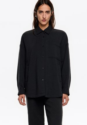 Camisa negra, ligera y texturizada, con cierre de botones en la parte delantera, mangas largas, cuello y bolsillo en el pecho. Combinada con pantalones a juego.