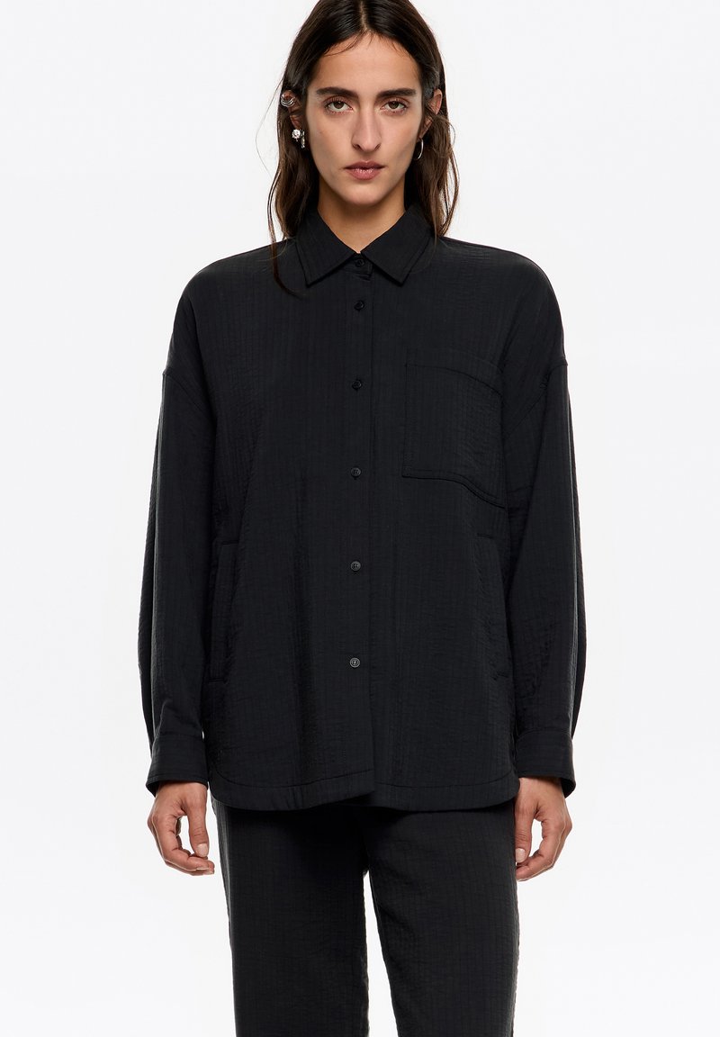 Camisa negra, ligera y texturizada, con cierre de botones en la parte delantera, mangas largas, cuello y bolsillo en el pecho. Combinada con pantalones a juego.