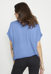 Blusa corta eampia a maniche corte di colore azzurro chiaro, in tessuto strutturato. Presenta un orlo arrotondato e spacchi laterali, abbinata a pantaloni neri.