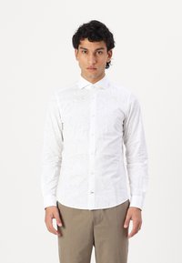 Cămașă albă cu mânecă lungă, cu un subtil model floral embossat, guler clasic și închidere cu nasturi, asortată cu pantaloni deschiși la culoare, maro.