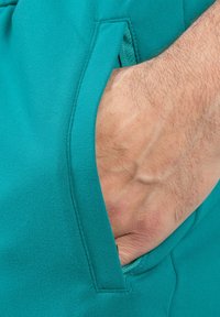 Tejido de chándal turquesa con una mano en un bolsillo lateral. El material parece suave con un ligero estiramiento; el bolsillo cuenta con un acabado de costura limpio.