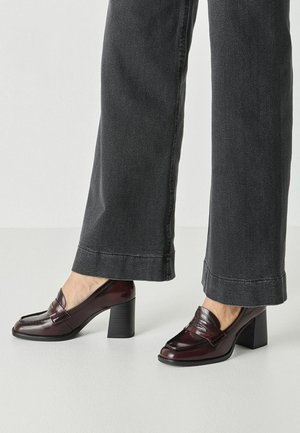 Jeans grigio scuro a gamba larga indossati con mocassini marrone scuro lucidi con tacco alto su pavimento chiaro e sfondo semplice.