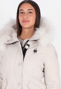 Chaqueta de invierno beige con capucha de piel sintética, cierre de botones, bolsillos laterales y un detalle de cremallera negra. Presenta un logo en la manga.