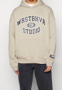 Beige hoodie met grote frontprint "WRSTBHVR STUDIO." Voorzien van een kangoeroezak en een zwart logolabel op de mouw. Zachte stof en een ontspannen pasvorm.