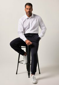 Biała koszula z długim rękawem i kieszenią zapinaną na guziki, w połączeniu z granatowymi joggerami i białymi sneakersami, siedząca na czarnym stołku na jasnym tle.