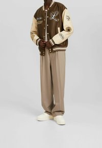 Brązowa kurtka typu varsity z kremowymi rękawami, ozdobiona naszywkami i haftowanymi literami, w połączeniu z beżowymi spodniami i białymi trampkami.