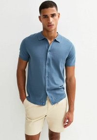 Camisa de manga corta, con botones, en un suave tejido de punto azul, que presenta un cuello clásico y una sutil textura acanalada, combinada con pantalones cortos beige.