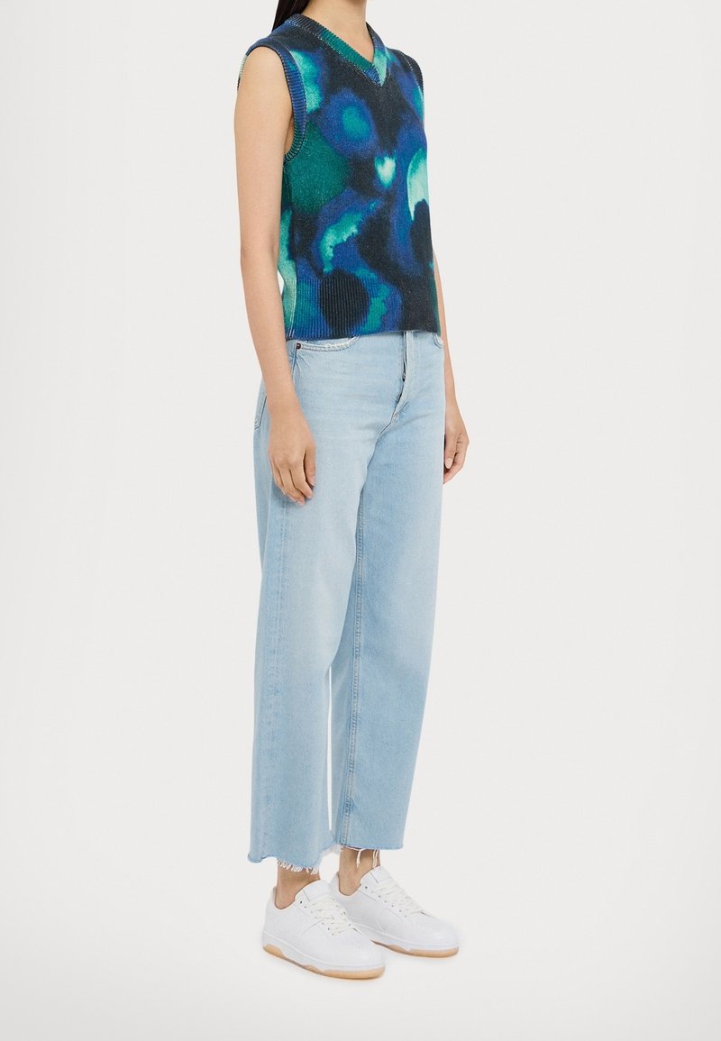Femme portant un pull sans manches bleu-vert à motifs, un jean court bleu clair aux ourlets effilochés et des baskets blanches à semelles en gomme.