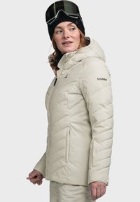 Hellbeige isolierte Jacke mit Kapuze, vertikalen Nähten und einem Logo auf dem Ärmel. Das Modell trägt Skibrillen und eine schwarze Mütze.