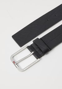 Cinturón de cuero negro con un acabado texturizado, que presenta una hebilla rectangular de metal plateado con grabado y un pequeño acento en rojo, blanco y azul.