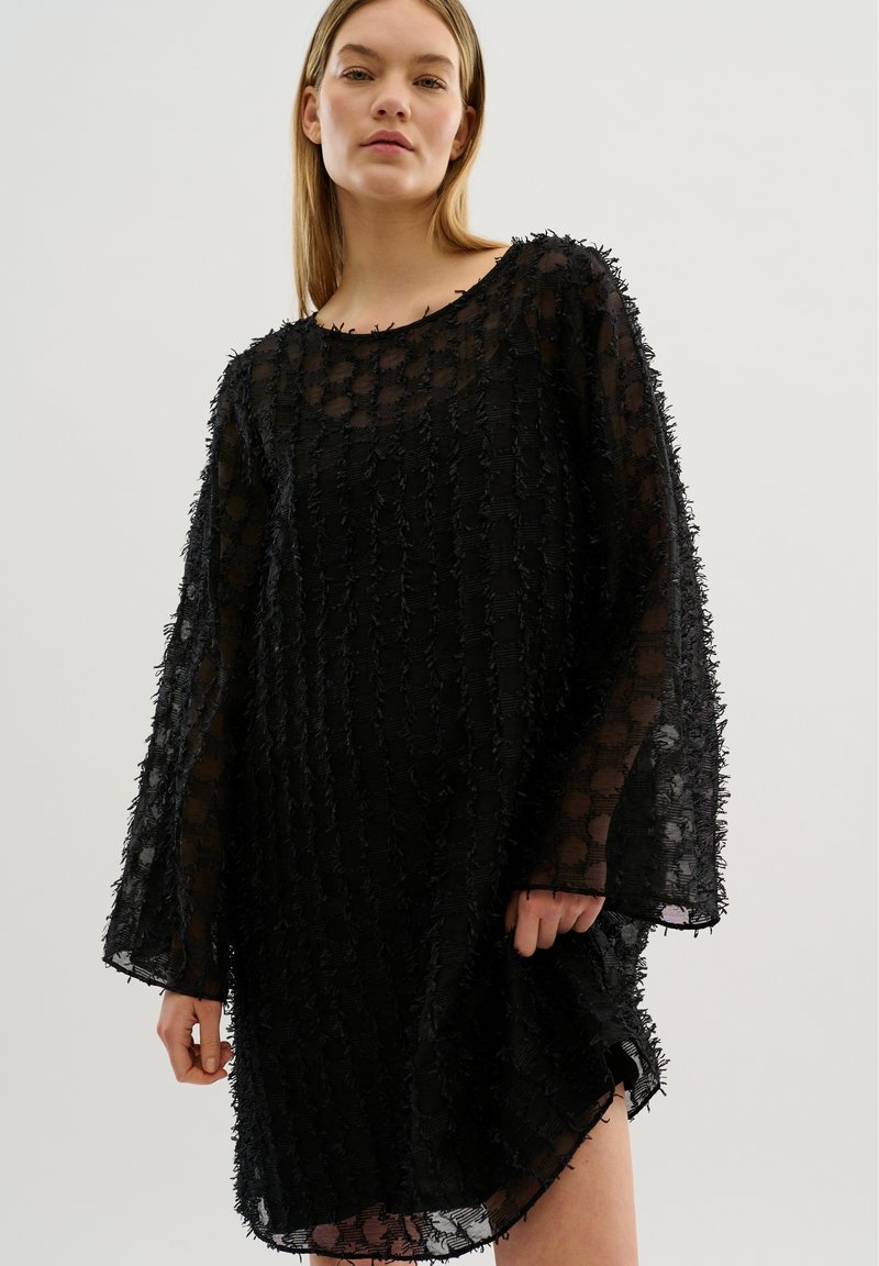 Black sheer dress featuring textured fabric with layered, ruffled accents, a loose fit, and wide sleeves. Hemline is slightly asymmetrical.