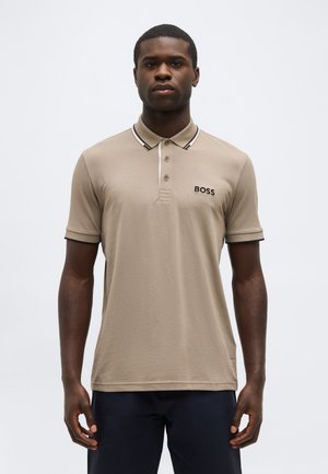 Mand iført beige poloshirt med sort og hvid kant på kraven og "BOSS" logo på brystet, stående mod en ensartet lys baggrund.