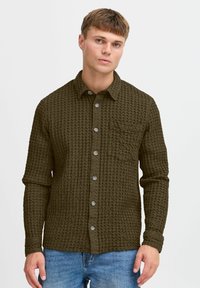 Camisa texturizada verde oliva con cuello abotonado, bolsillo frontal y botones visibles, combinada con jeans de mezclilla azul.