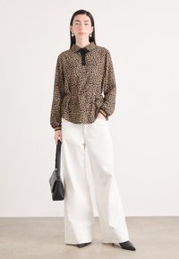 Blouse met luipaardprint in bruintinten, knoopsluiting met zwarte kraag en strik, gecombineerd met wijde witte broek en puntige zwarte hakken.