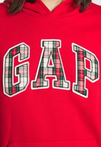 Červená mikina s kapucí s velkými aplikovanými písmeny "GAP" v kárované látce s bílým prošíváním. Měkký materiál s kapsou klokankou.