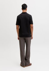 Man staand met de rug naar de kijker gekeerd, draagt een zwart poloshirt met korte mouwen, grijze broek, bruine schoenen en een zilveren armband aan de linkerpols.