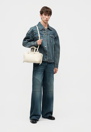 Vīrietis valkā zilu džinsa jaku un platajiem galiem džinsus, turot baltu somiņu ar plecu siksnu uz vienkrāsaina fona.