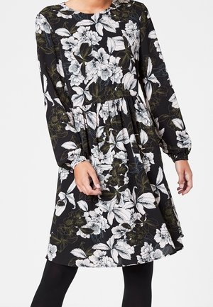 Musta pitkähihainen mekko, jossa valkoinen ja vihreä kukkakuvio, vyötäröllä laskokset ja hieman levenevä helma.