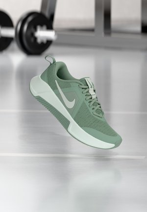 Zielony but sportowy Nike z białym logo Swoosh, unoszący się nad błyszczącą podłogą siłowni, z rozmytymi ciężarkami w tle.