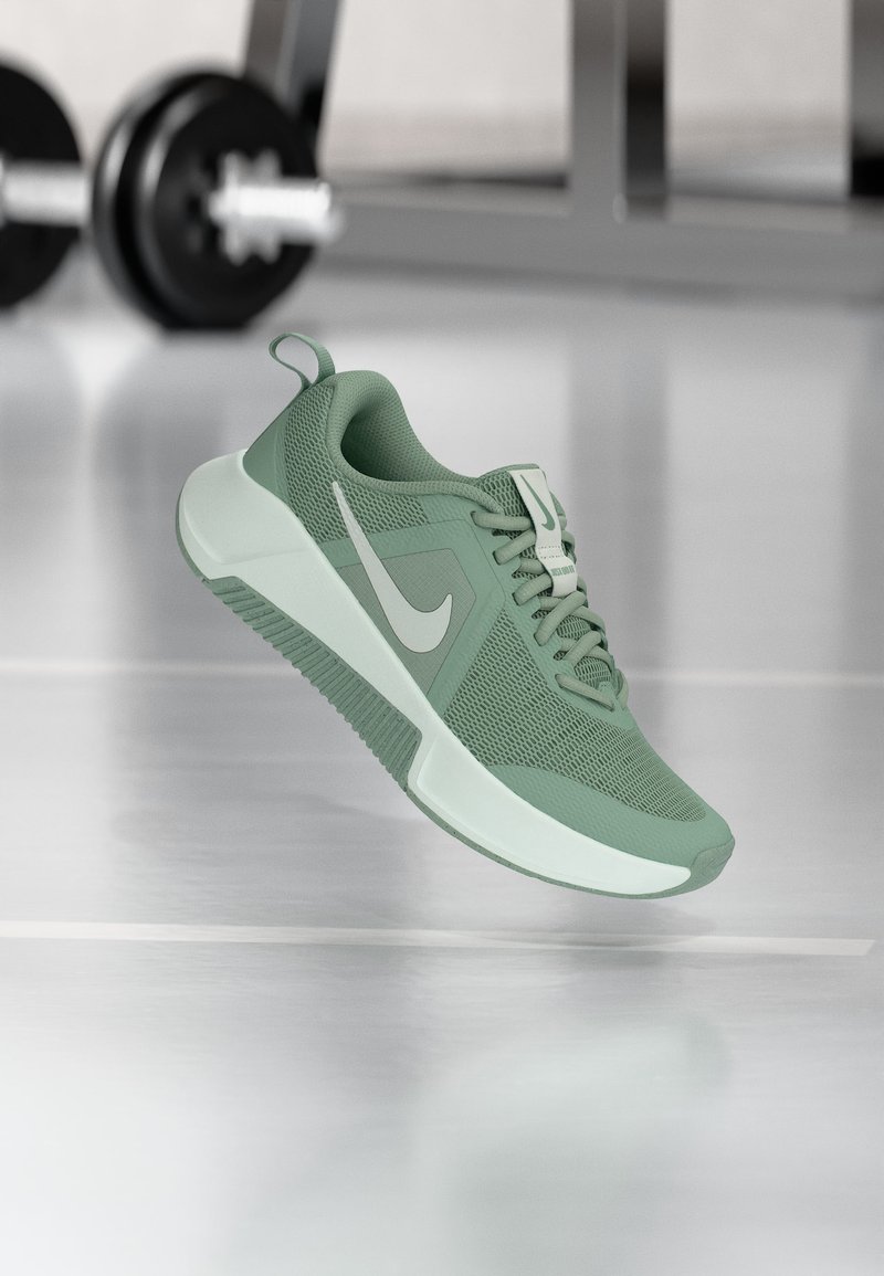 Πράσινο αθλητικό παπούτσι Nike με λευκό λογότυπο σβούρας, αιωρούμενο πάνω από γυαλιστερό δάπεδο γυμναστηρίου με φόντο ασαφή βάρη.