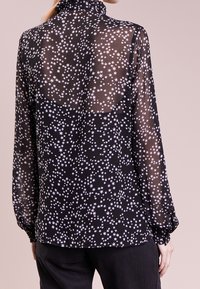 Blouse noire transparente à manches longues avec motif d'étoiles blanches portée avec un pantalon noir, vue de dos.