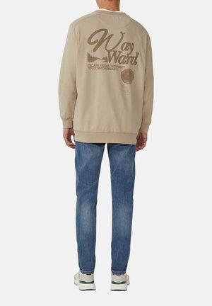 Beige sweatshirt met contrasterende bruine grafische tekst op de achterkant. Lichtblauwe spijkerbroek en witte sneakers met groene accenten. Casual fit.