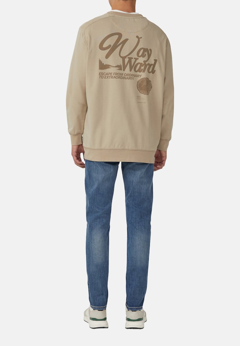 Beige sweatshirt met contrasterende bruine grafische tekst op de achterkant. Lichtblauwe spijkerbroek en witte sneakers met groene accenten. Casual fit.