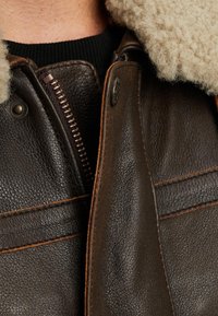 Brun läderjacka med fårskinnskrage, en koppardragkedja, kontrasterande sömmar, en strukturerad yta och knappdetaljer.