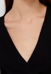 Haut cache-cœur texturé noir avec un col en V profond, présentant un tissu doux et un motif subtil ; vue rapprochée mettant en valeur le col et la région des épaules.