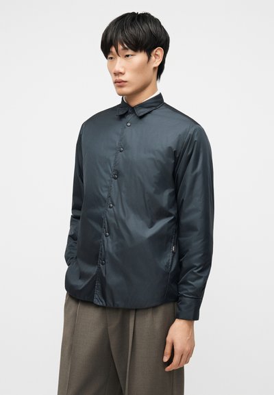 Άνδρας που φοράει σκοτεινό ναυτικό κουμπωτό σακάκι με γιακά και γκρι παντελόνι με φαρδιά πόδια, στέκεται μπροστά σε απλό φωτεινό φόντο.