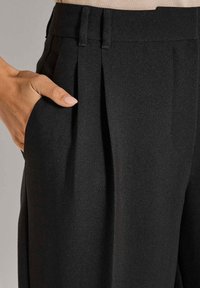 Hand rustend in de zak van hoge taille, zwarte plooibroek met riemlussen en op maat gemaakte naden.