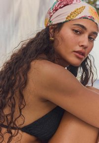 Zwarte textured bikini top, hoofddoek met bloemenpatroon en gouden hoepeloorbel; krullend haar dat over de schouder valt, natuurlijke huidskleur en uitstraling.