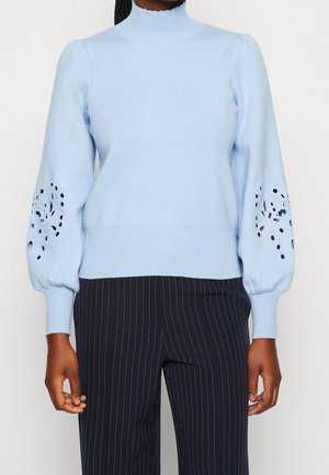 Femme portant un pull bleu clair avec des découpes florales sur des manches bouffantes et un pantalon sombre à fines rayures, debout devant un fond blanc.