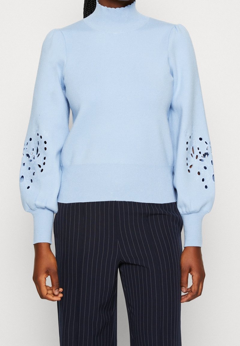 Femme portant un pull bleu clair avec des découpes florales sur des manches bouffantes et un pantalon sombre à fines rayures, debout devant un fond blanc.