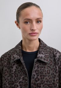 Woman with slicked-back hair, wearing small hoop earrings, a leopard print jacket with a zipper, and a black ribbed top.