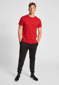 Rød kortærmet t-shirt parret med sorte sweatpants, der har elastiske manchetter og lommer, båret med sorte sportssko på en ensfarvet baggrund.