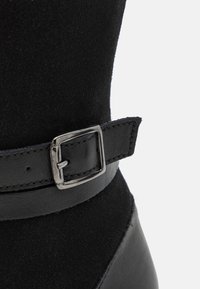 Bottine noire avec un dessus en daim doux, une lanière en cuir brillant et un détail de boucle en argent, soulignant une silhouette structurée.