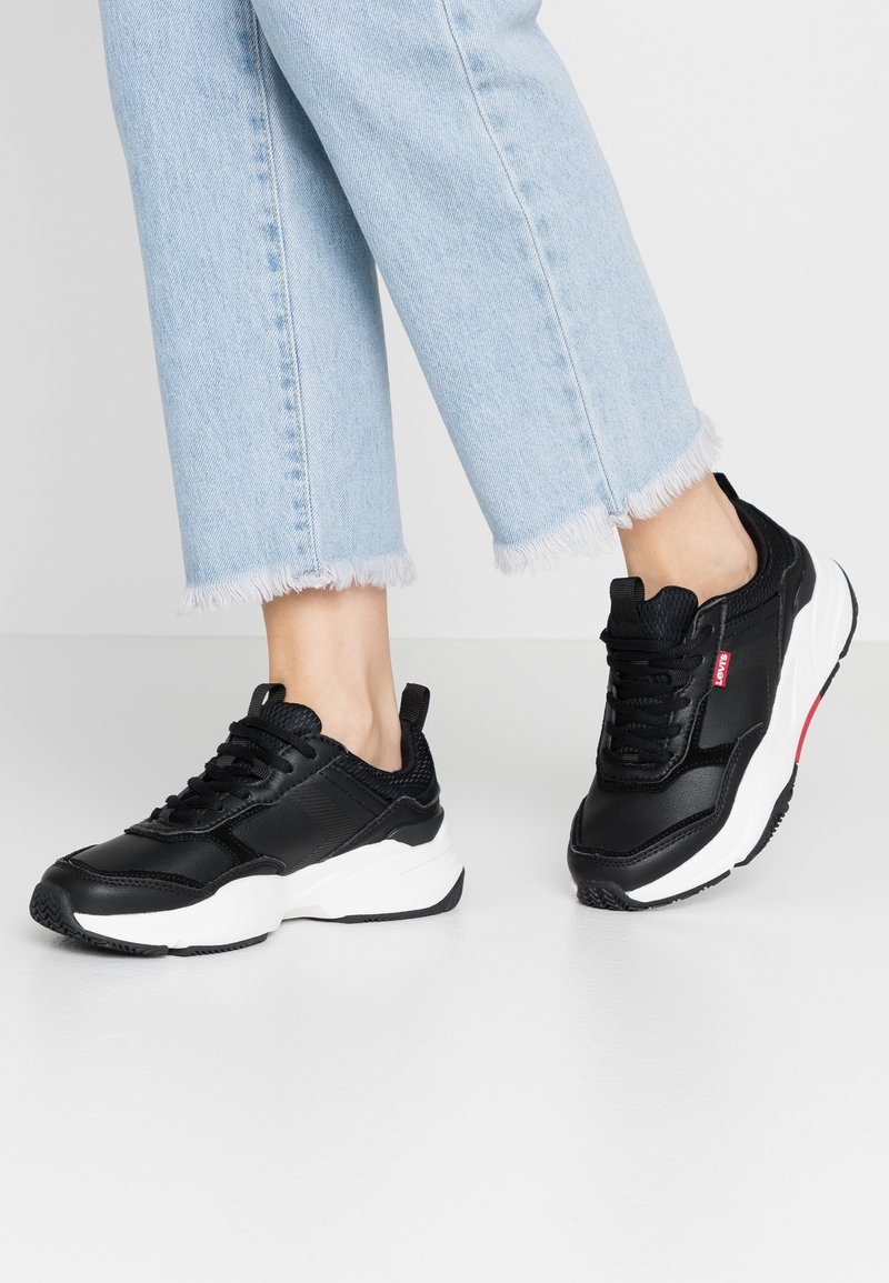 Černé sportovní boty s texturovaným povrchem, bílou podrážkou a červeným akcentem. Design obsahuje kulaté tkaničky a poutko na patě.