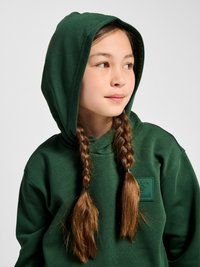 Jeune fille avec des cheveux bruns tressés portant un sweat à capuche vert foncé, regardant vers la droite sur un fond clair uni.