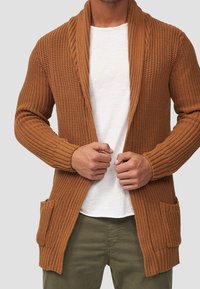 Brązowy dzianinowy kardigan z kołnierzem typu szal, prążkowanymi mankietami i dwiema przednimi kieszeniami. Noszony na białej koszuli z textured designem.