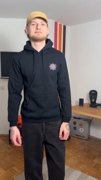 Μαύρο φούτερ με κουκούλα, με γραφικό λογότυπο στο στήθος, κουκούλα με κορδόνι και τσέπη καγκουρό, σε συνδυασμό με σκούρο παντελόνι και μπεζ καπέλο.