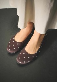 Braune Ballettflachetten mit Polka-Dot-Muster, glatter Textur und abgerundetem Zehenbereich, kombiniert mit einem cremefarbenen, fließenden Kleid, auf einer dunklen Oberfläche.