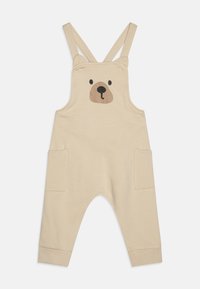 Beige overall med bjørneansigt-design på brystet, med justerbare stropper, sidelommer og manchetter ved anklerne for komfort. Glat tekstur.