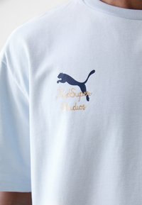 Gaiši zila kokvilnas t-kreklu ar tumši zilu Puma logotipu un zelta "KidSuper Studios" tekstu izšūtu uz krūts. Īsas piedurknes, apaļa kakla izgriezums.