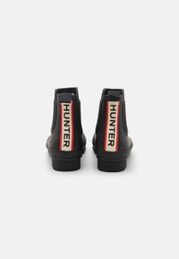 Μαύρες καουτσούκ μπότες έως τον αστράγαλο με μαλακή υφή, ψηλό τακούνι, πίσω λωρίδες και έντονο κόκκινο και λευκό λογότυπο "HUNTER" στο πίσω μέρος.