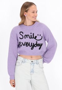 Haken trui in lavendel met zwarte tekst "Smile everyday", geribbelde zoom en lange mouwen. Gemaakt in combinatie met licht gewassen, high-waisted spijkerbroek.