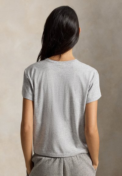 Kobieta o długich ciemnych włosach, ubrana w jasnoszary t-shirt z krótkim rękawem i pasujące szare spodnie, stojąca z rękami w kieszeniach, zwrócona tyłem.