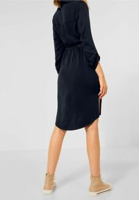 Svart klänning med långa ärmar, bältad midja, sprund i sidorna och rundad fåll. Kombinerad med beige ankelstövlar mot en neutral bakgrund.