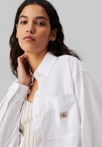 Hvid skjorte med knapper og lomme, der har et CK-logo-patch. Stoffet ser glat ud med en struktureret design. Modellen poser blidt.