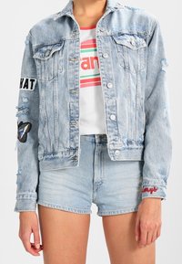 Giacca in denim azzurro chiaro con finitura sbiadita, chiusura a bottoni, tasche frontali e toppe ricamate a forma di farfalla e con testo sulle maniche.